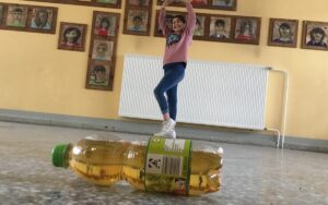 Im Vordergrund liegt eine Apfelschorleflasche auf dem Boden und im Hintergrund ist ein Mädchen. Die Perspektive des Fotos wurde so gewählt, dass es den Anschein erweckt, dass das Mädchen auf der viel zu großen Flaschen steht.