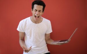 Ein Junger Mann steht vor einer rotten Wand. Er schreit und hält in seiner Hand ein Notebook