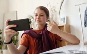Ein junges Mädchen sitzt am Schreibtisch und macht ein Selfie mit ihrem Handy und sie lacht dabei