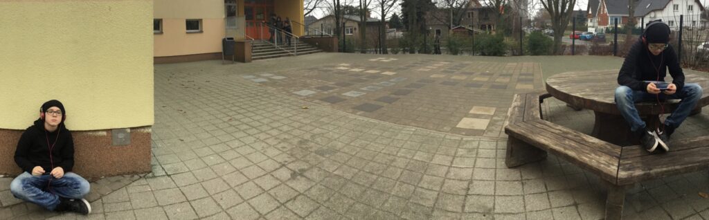 Ein bearbeitets Foto mit einem Jungen in zwei verschiedenen Positionen auf einem Schulhof. Auf der rechten Seite des Fotos sitzt er auf einem Tisch und spielt mit seinem Smartphone. Auf der linken Seite sitzt er auf dem Boden und hört Musik mit Kopfhörer