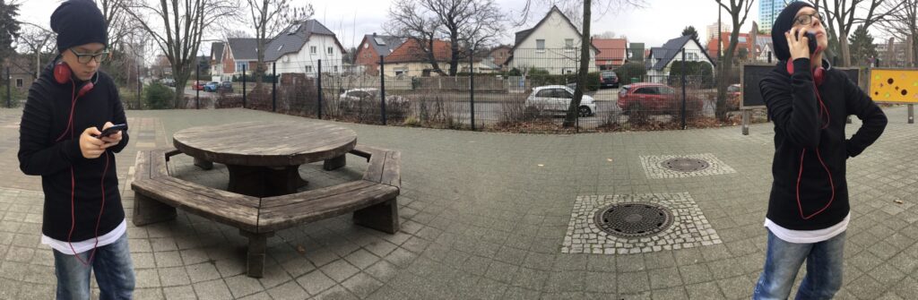 Ein bearbeitets Foto mit einem Jungen in zwei verschiedenen Positionen auf einem Schulhof. Auf der rechten Seite des Fotos steht er und telefoniert mit seinem Smartphone. Auf der linken Seite tippt er mit dem Smartphone.