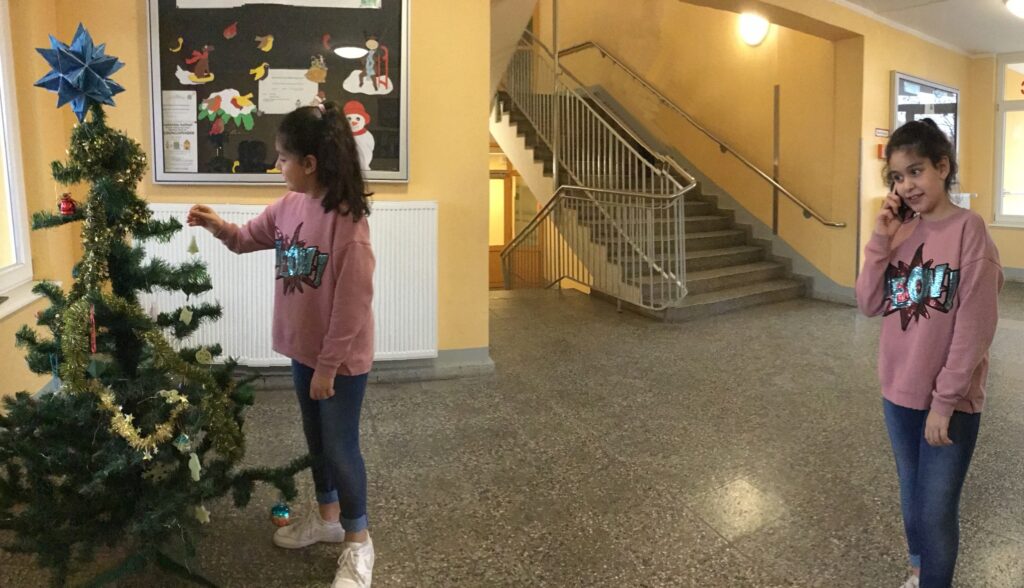 Ein bearbeitets Foto mit einem Mädchen in zwei verschiedenen Positionen. Auf der rechten Seite steht sie und telefoniert mit dem Smartphone. Auf der linken Seite steht sie neben einem geschmückten Weihnachtsbaum.