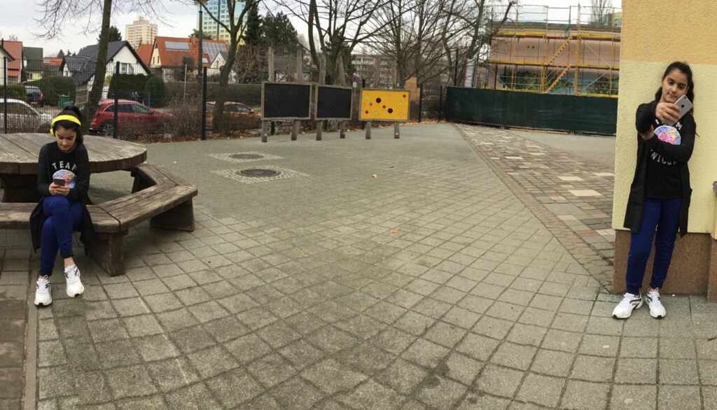 Ein bearbeitets Foto mit einem jungen Mädchen Auf dem Schulhof in zwei verschiedenen Positionen. Auf der rechten Seite macht sie ein Selfie mit ihrem Smartphone. Auf der linken Seite sitzt sie auf einer Bank und schaut auf das Smartphone-Display während sie Musik mit Kopfhörern hört.