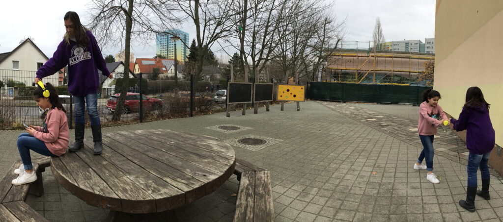 Ein bearbeitets Foto mit zwei jungen Mädchen Auf dem Schulhof in zwei verschiedenen Positionen. Auf der rechten Seite des Fotos streiten sie um Kopfhörer. Auf der linken Seite versucht das eine Mädchen die Kopfhörer von dem anderen wegzunehmen.