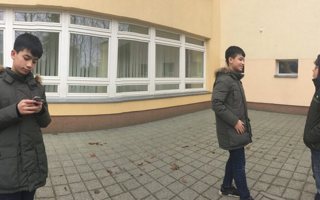 Ein bearbeitets Foto mit einem Jungen in zwei verschiedenen Positionen auf einem Platz. Auf der rechten Seite spricht mit einem anderen Jungen. Auf der linken Seite tippt er mit dem Handy