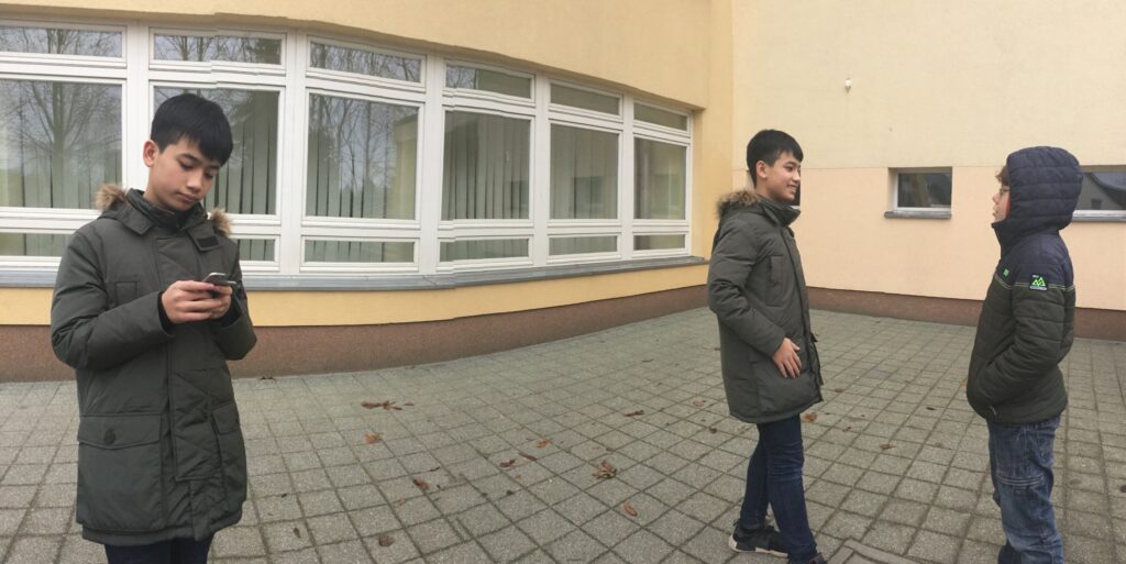 Man sieht drei Kinder im Bild. Einer von denen schaut ins Handy und die anderen zwei reden miteinander.