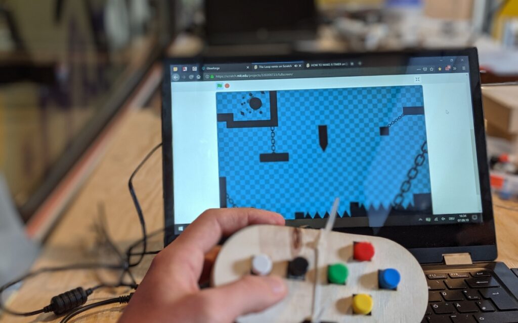 Im Vordergrund hält eine Hand eine hölzerne, handgefertigte Steuerung und im Hintergrund liegt ein Notebook auf dem Tisch. Auf dem Bildschirm läuft ein Spiel.