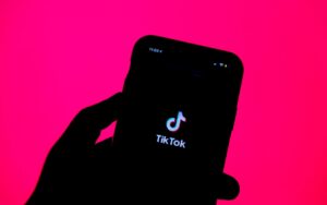 Eine Hand hält ein Smartphone vor einer rosa Wand. Auf dem Display des Smartphones ist das Logo der App TikTok zu sehen.