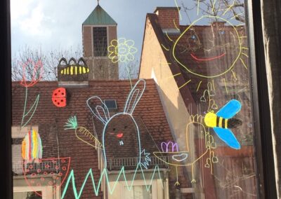 Ein Fensterglas, auf dem Dinge wie Sonne, Blumen, eine Biene mit Farben gezeichnet wurden. Hinter dem Fensterglas ist ein Haus und ein Kirchturm zu sehen.