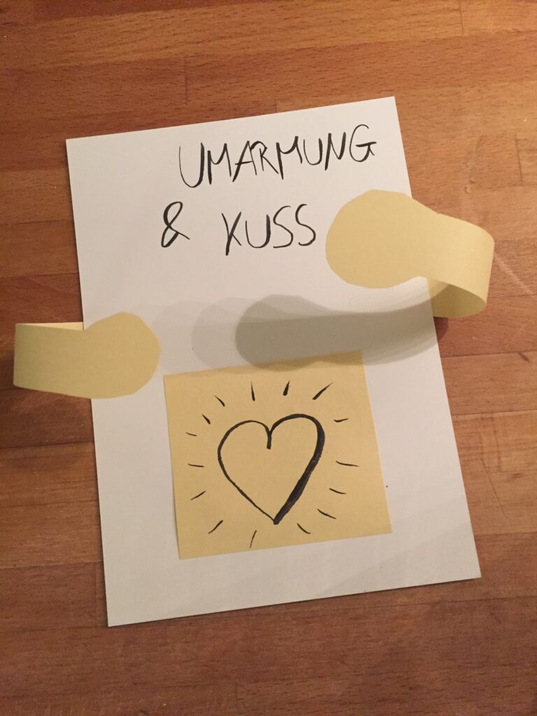 Eine Karte mit der Aufschrift "Umarmung & Kuss". Aus der Karte kommen zwei Arme raus. In der Mitte der Karte ist ein Herz: