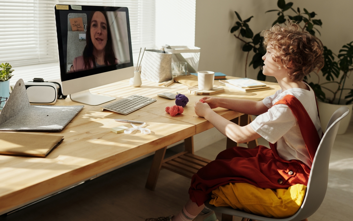 Ein Kind sitzt am Tisch vor einem Rechner und hat einen Video Anruf mit einer Frau