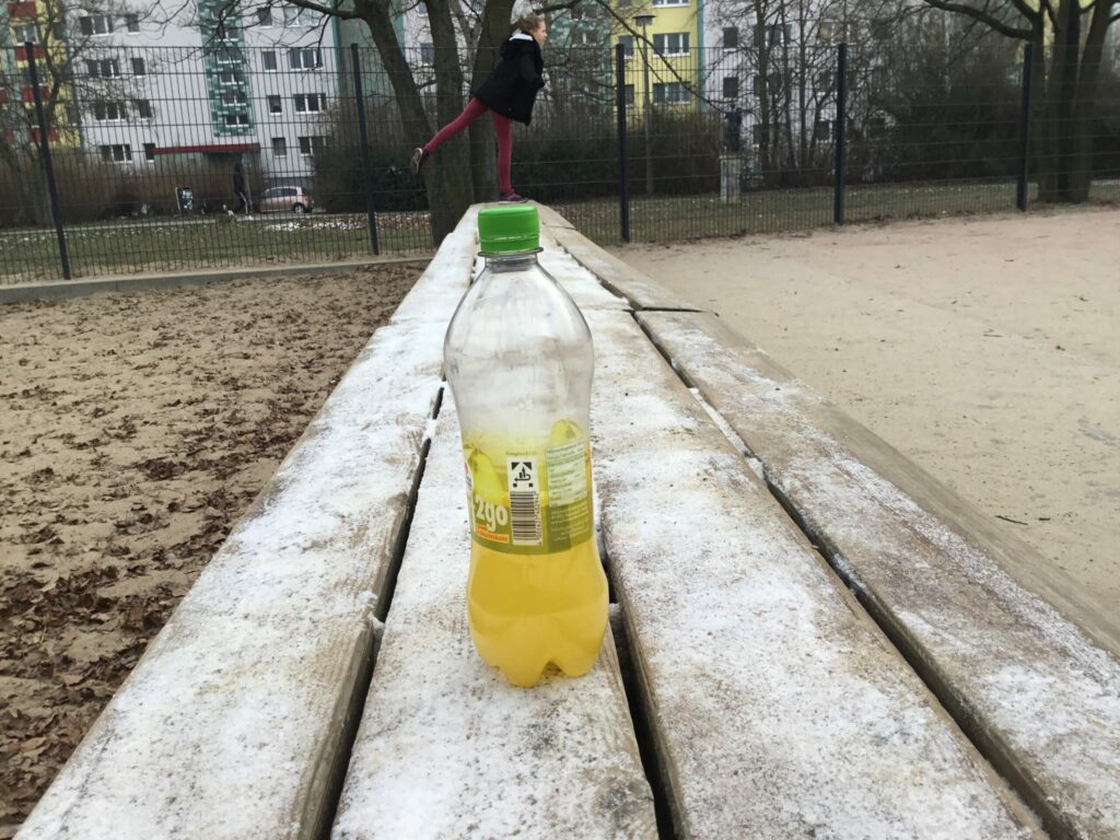 Im Vordergrund des Fotos steht eine Saftflasche auf dem Tisch und im Hintergrund ein Mädchen. Die Perspektive des Fotos wurde so gewählt, dass es den Anschein erweckt, dass das Mädchen nur mit einem Fuß auf der Saftflasche balanciert