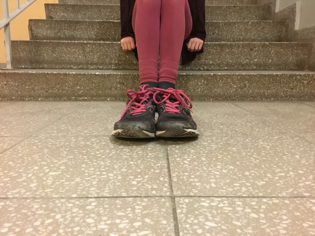 Ein Mädchen sitzt auf den Treppen. Im Vordergrund stehen Schuche durch eine optische Täuschung sehen sie jedoch größer aus.