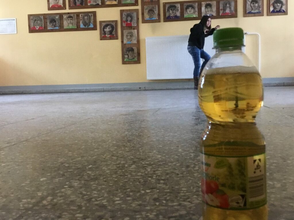 Im Vordergrund steht eine Apfelschorleflasche auf dem Boden und im Hintergrund ist ein Mädchen. Die Perspektive des Fotos wurde so gewählt, dass es den Anschein erweckt, dass das Mädchen auf die Apfelschorleflasche hinaufklettert.