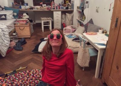 Eine junge Frau sitzt in ihrem Zimmer auf dem Boden. Sie trägt eine Brille in Form von zwei Erdbeeren.