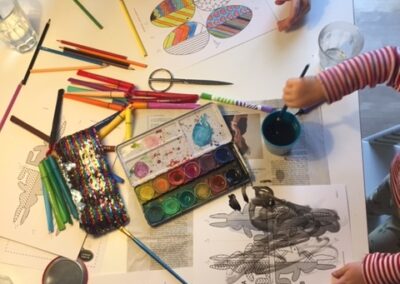 Zwei Kinder sitzen an einem Tisch und malen mit Farben. Auf dem Tisch liegen viele bemalte Blätter sowie Bleistiftfarben und Wasserfarben.
