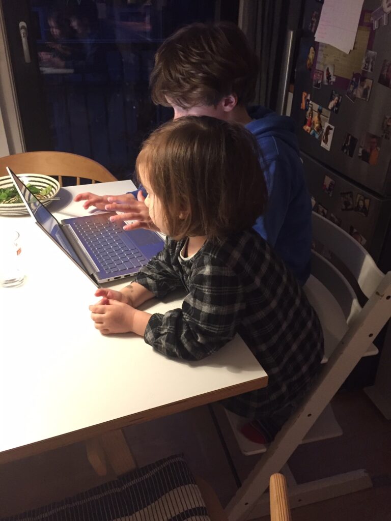 Ein kleiner Junge sitzt am Esstisch vor einem Notebook in der Küche. Neben ihm sitzt ein kleines Mädchen.