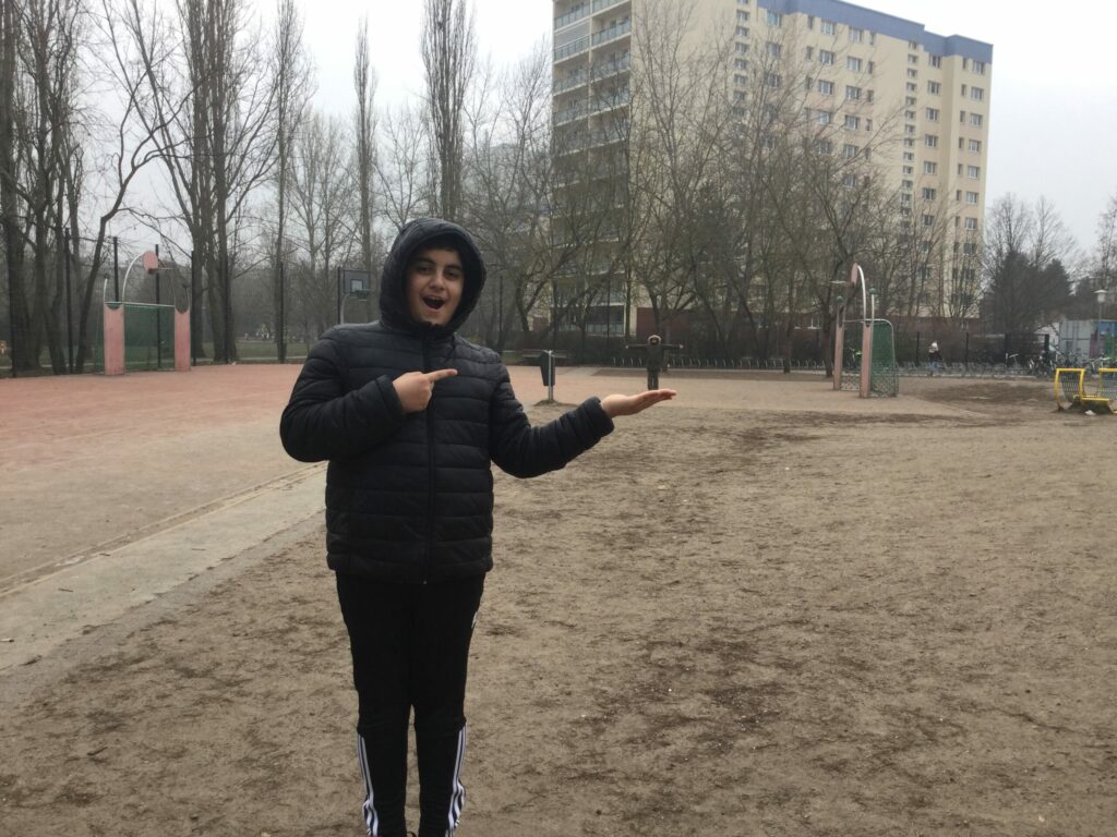 Im Vordergrund des Fotos steht ein Junge und im Hintergrund ganz klein ein anderes Kind. Die Perspektive des Fotos wurde so gewählt, dass es den Anschein erweckt, dass der Junge das Kind auf seiner Hand hält.