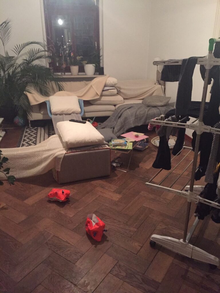 Ein Wohnzimmer. Im Hintergrund steht ein Sofa und im Vordergrund ein Wäscheständer. Auf dem Boden liegen Kinderspiele.