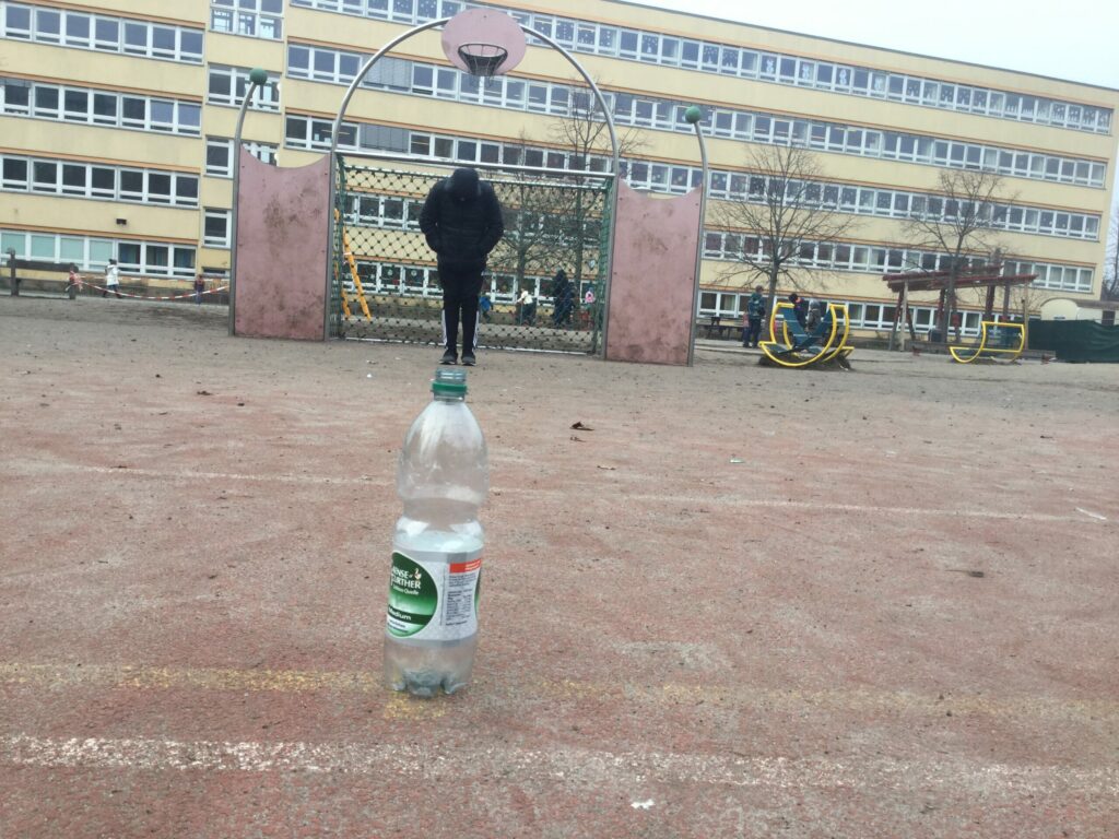 Im Vordergrund steht eine Wasserflasche auf dem Boden und im Hintergrund ist ein Junge. Die Perspektive des Fotos wurde so gewählt, dass es den Anschein erweckt, dass der Junge auf der viel zu großen Flaschen steht.