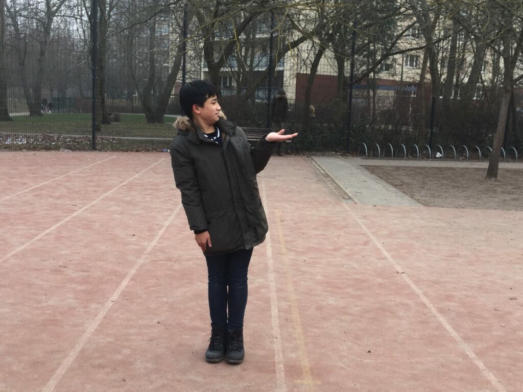 Im Vordergrund steht ein Junge, der seine Hand ausstreckt. Im Hintergrund steht eine Person auf der Bank. Die Perspektive des Fotos wurde so gewählt, dass es den Anschein erweckt, dass die Person im Hintergrund auf der Hand des Jungen im Vordergrundsteht