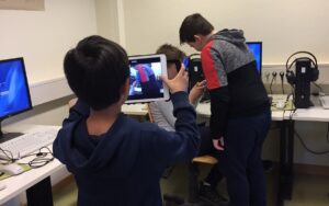 Drei Jungs sitzen in einem Computerraum. Einer von ihnen nimmt ein Video von den anderen zwei auf.