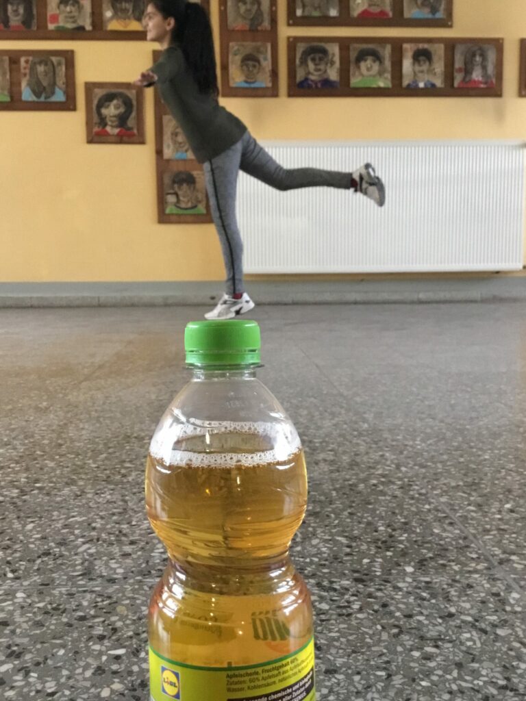 Im Vordergrund des Fotos steht eine Saftflasche auf dem Tisch und im Hintergrund ein Mädchen. Die Perspektive des Fotos wurde so gewählt, dass es den Anschein erweckt, dass das Mädchen nur mit einem Fuß auf der Saftflasche balanciert