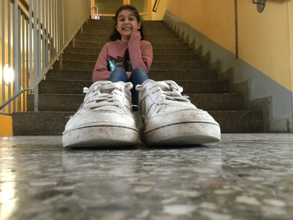 Im Vordergrund sieht man Schuhe und ihm Hintergrund sieht man ein lächelndes Mädchen