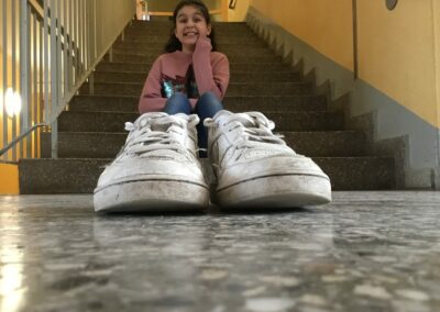 Im Vordergrund sieht man Schuhe und ihm Hintergrund sieht man ein lächelndes Mädchen