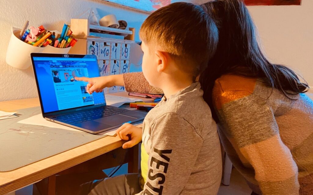 Ein kleines Kind sitzt am Schreibtisch vor einem Notebook. Neben ihm steht ein kleines Mädchen und zeigt mit ihrem Finger auf das Notebook-Display