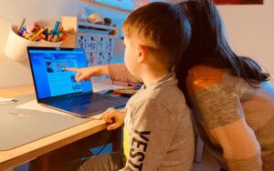 Ein kleines Kind sitzt am Schreibtisch vor einem Notebook. Neben ihm steht ein kleines Mädchen und zeigt mit ihrem Finger auf das Notebook-Display