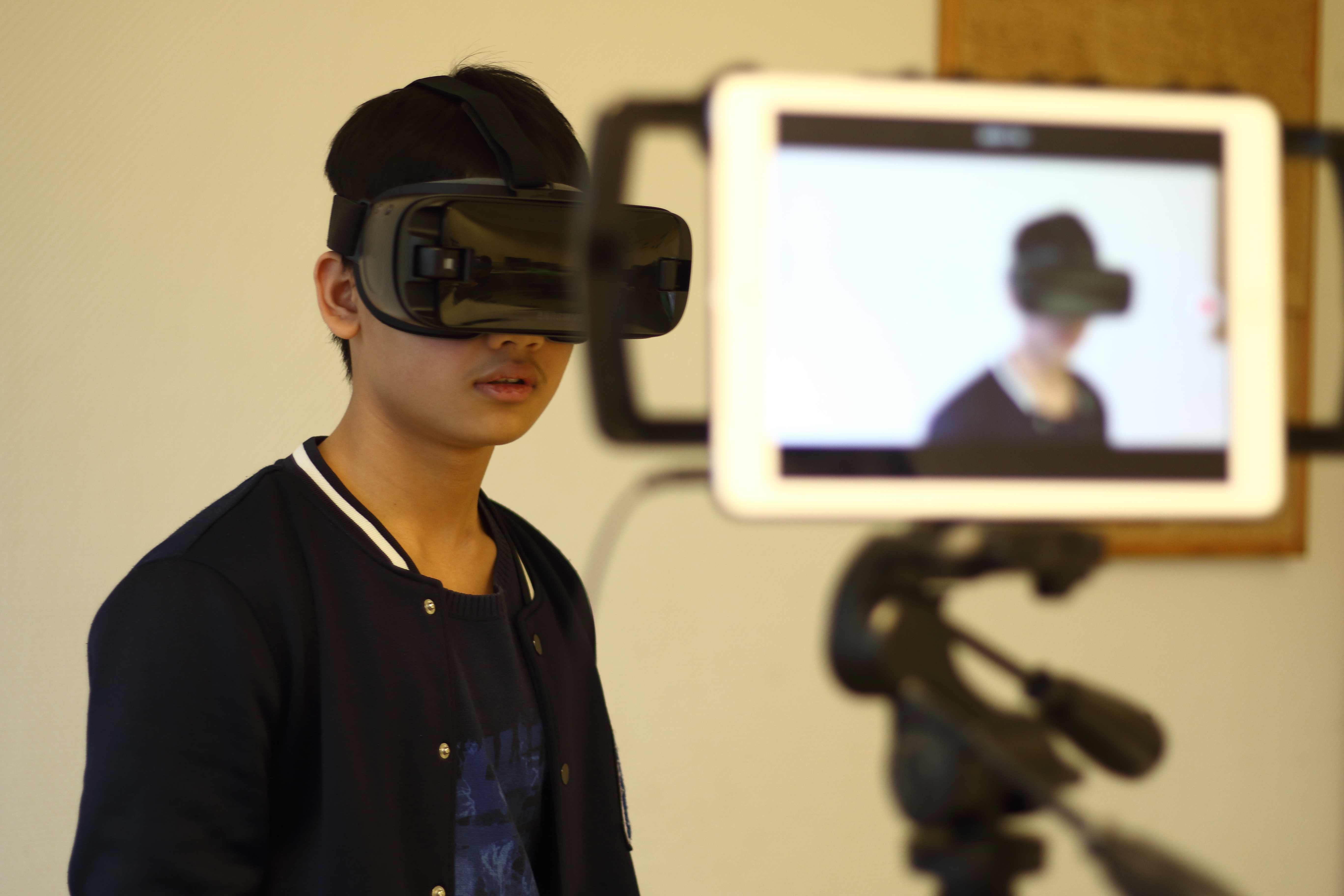 Ein Junge trägt eine VR Brille, gleichzeitig wird er von einem Tablet fotografiert beziehungsweis wird ein Video von ihm aufgenommen.