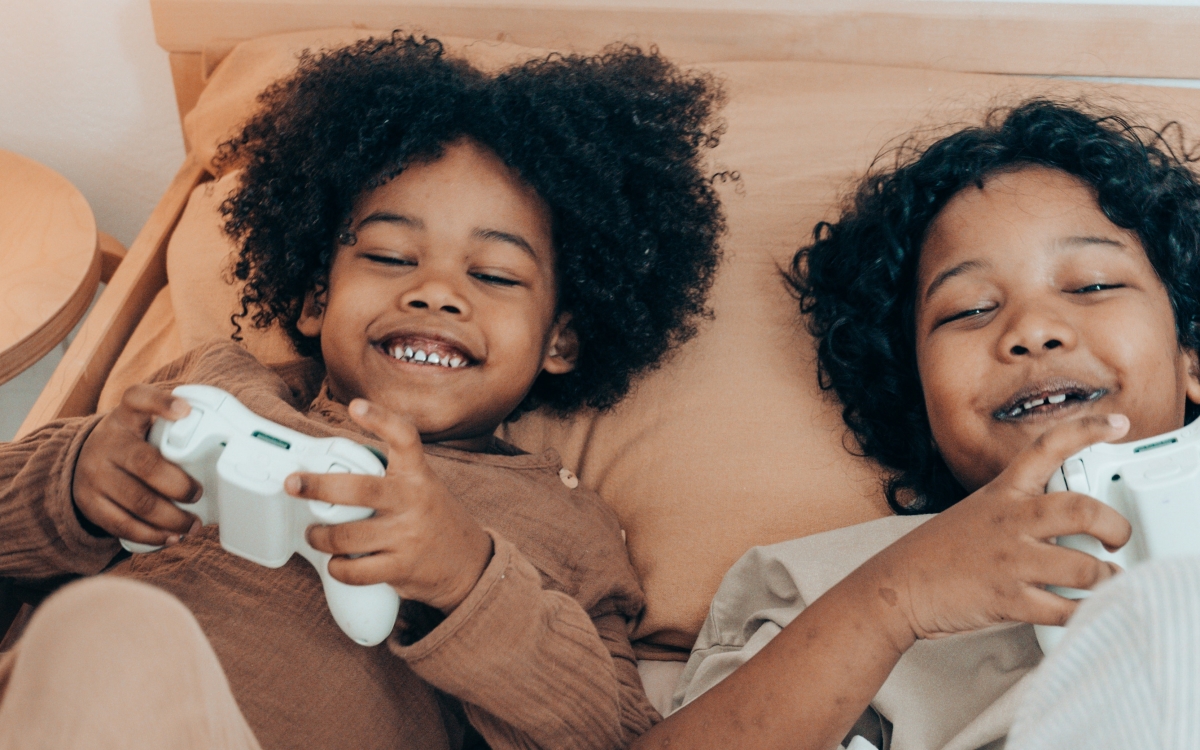 Ein kleiner Junge und ein kleines Mädchen spielen im Bett mit dem Kontroller einer Konsole und lachen dabei.