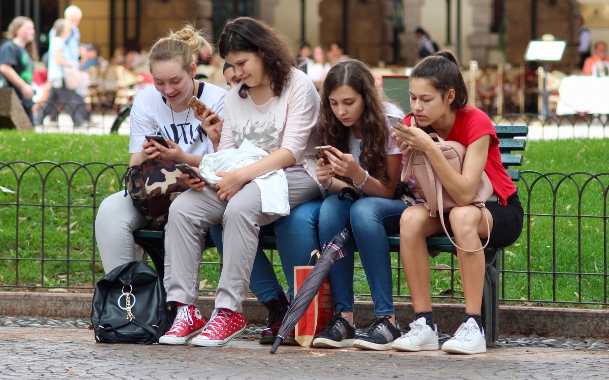 Fünf Mädchen sitzen nebeneinander auf einer Bank auf der Straße und jede schaut auf ihr Smartphone-Display.