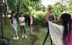 Im Hintergrund stehen vier junge Mädchen. Zwei von ihnen halten ein Mikrofon in der Hand. Eine andere steht im Vordergrund und nimmt ein Video von ihnen mit einem Ipad auf.