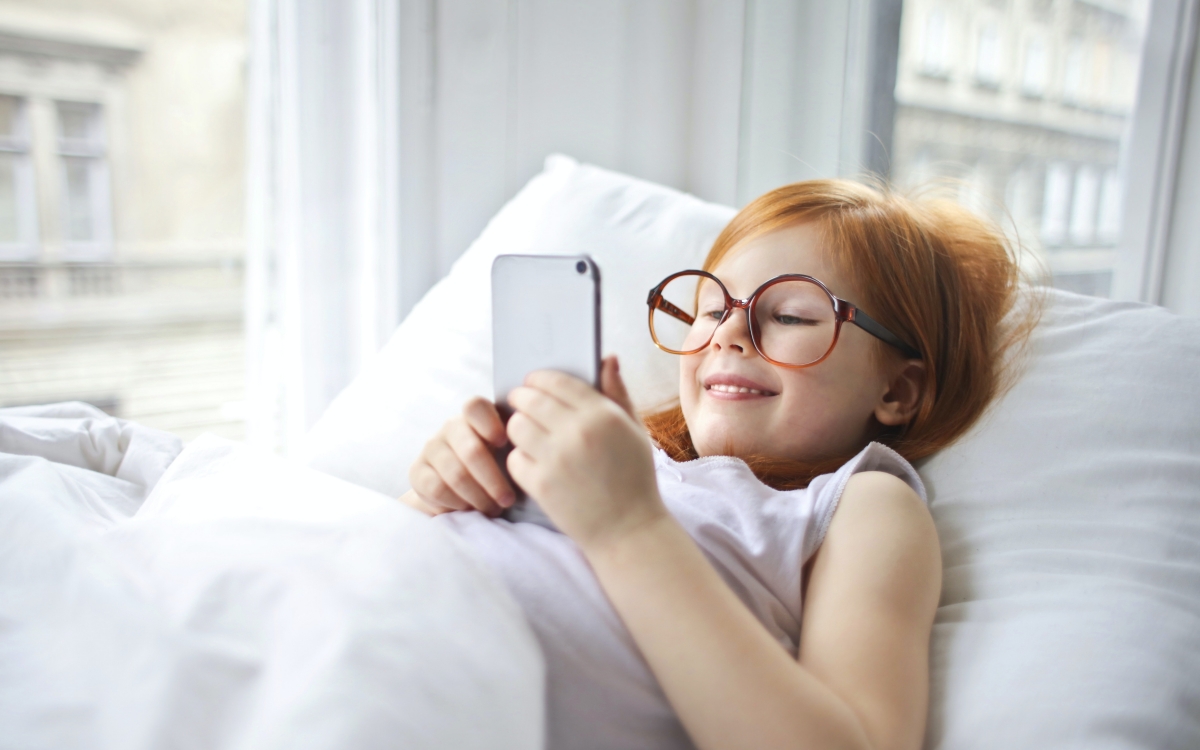 Ein Mädchen mit Brille liegt im Bett, während sie lächelnd in ihren Smartphone schaut