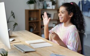Ein junges Mädchen in sitzt am Tisch und winkt in den Computer