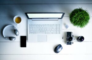 Ein Schreibtisch, der von oben aufgenommen wurde. Auf ihm befinden sich ein Notebook, eine Kamera, ein Handy, eine Maus, Kopfhörer, eine Kaffetasse und eine Pflanze.