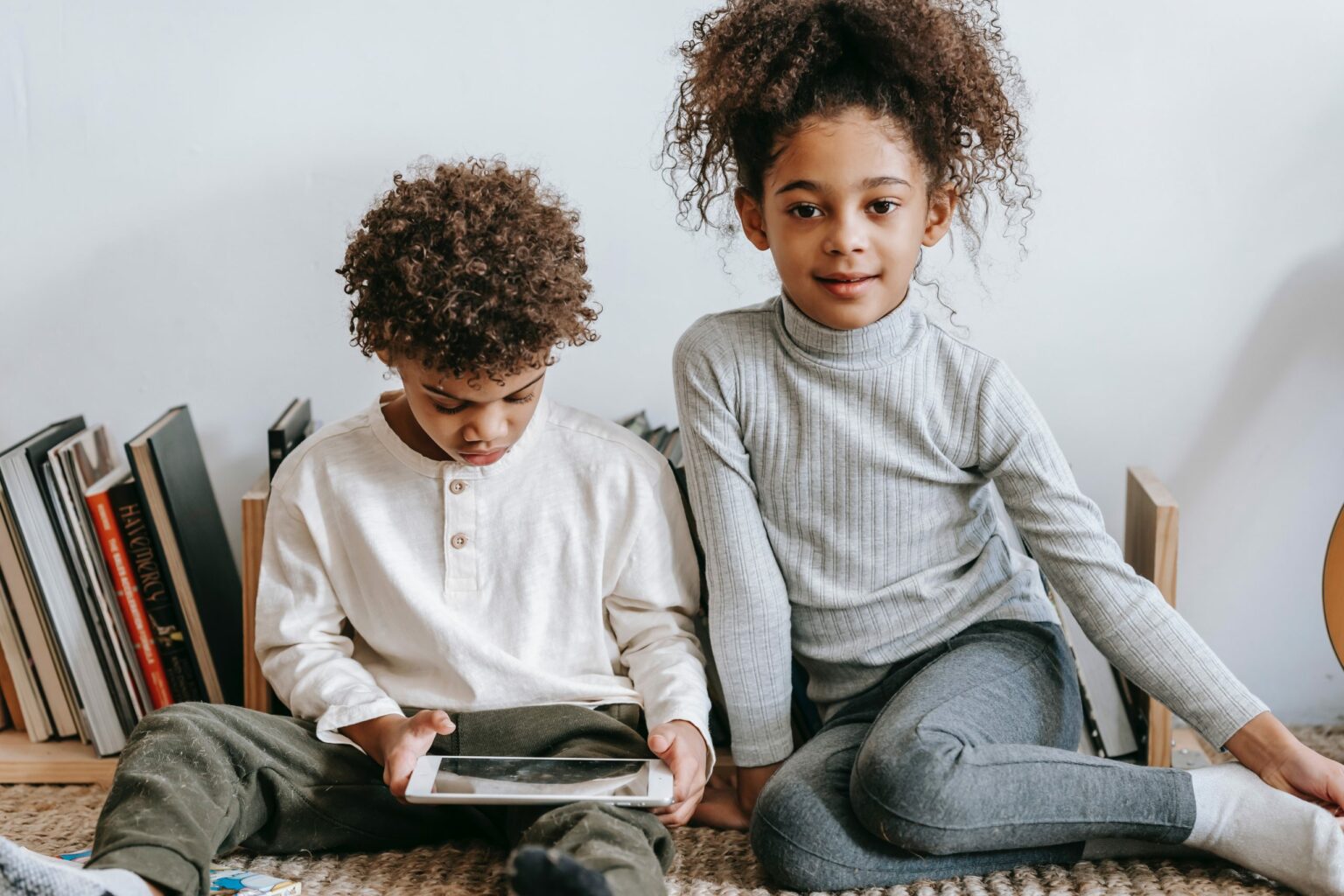 Zwei Kinder sitzen nebeneinander auf dem Boden. Der kleine Junge hält ein Tablet in den Händen, während das kleine Mädchen lächelnd in die Kamera schaut.