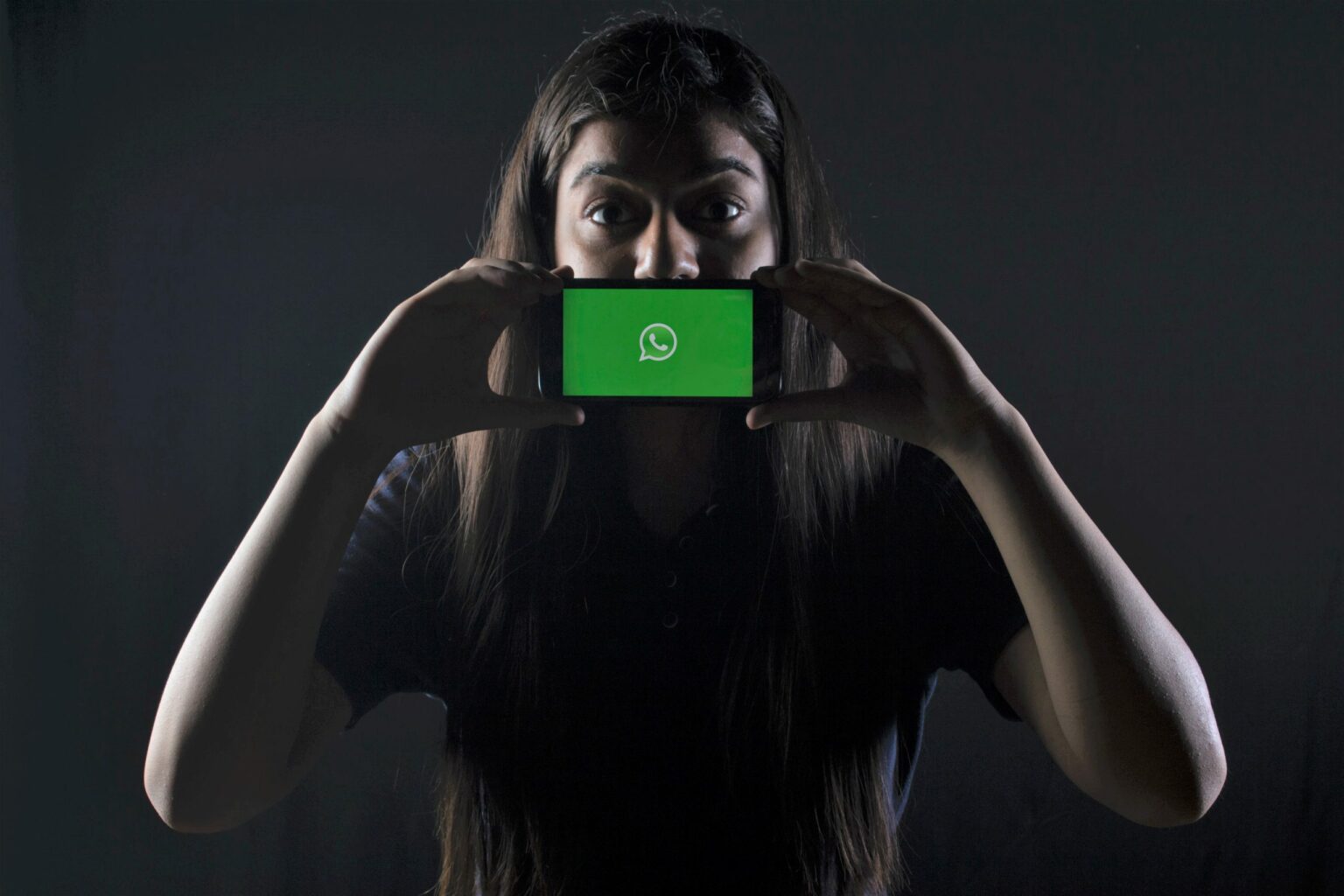 Ein Mädchen im Schulalter steht im Dunkeln und hält sich ihr Smartphone vor den Mund. Auf dem Display des Smartphones ist die App Whatsapp zu sehen.