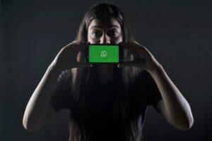 Ein Mädchen im Schulalter steht im Dunkeln und hält sich ihr Smartphone vor den Mund. Auf dem Display des Smartphones ist die App Whatsapp zu sehen.