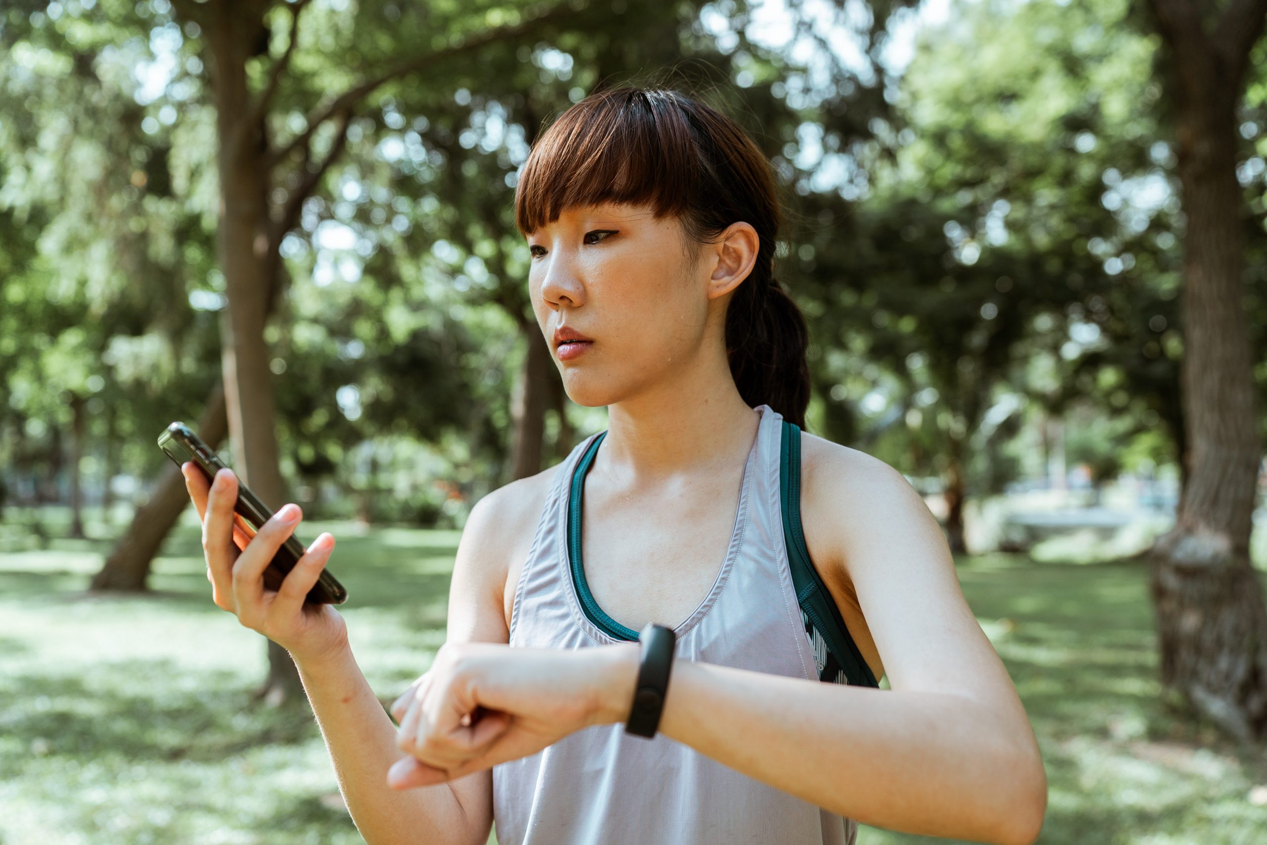 Eine Frau in Sportkleidung befindet sich in einem Park. Sie schaut auf ihr Smartphone und trägt ein Fitnessarmband.